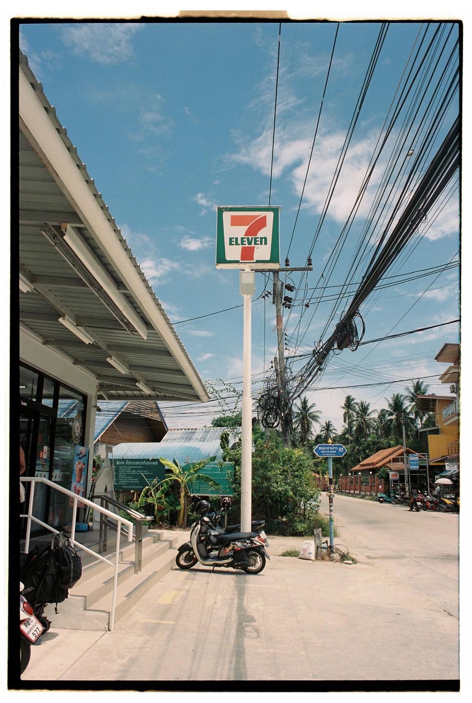 A 7-Eleven sign stands tall next to a building with a scooter parked outside, surrounded by palm trees and a street with many electrical wires overhead.