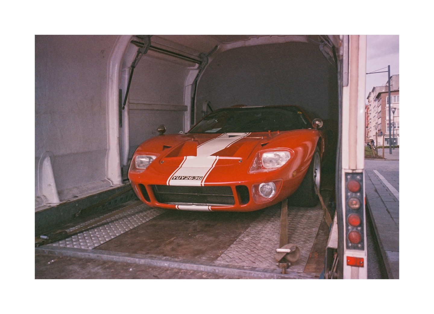 A red sports car with white racing stripes is parked inside the back of a truck.
