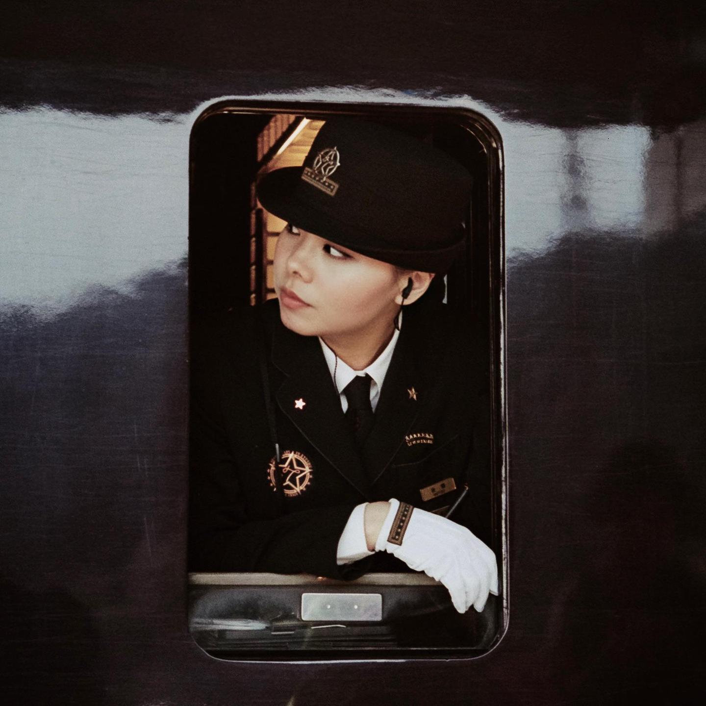 A woman in a uniform with a hat and white gloves looks out of a train window.