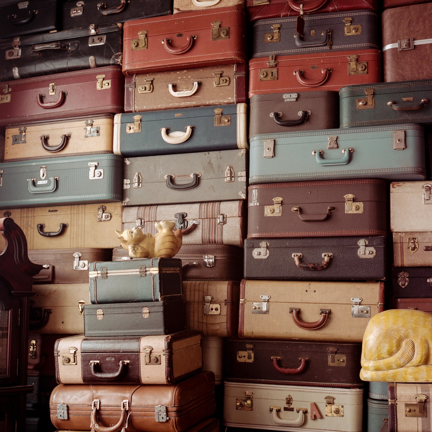 A large stack of vintage suitcases in various colors and sizes fills the frame, with a small ceramic elephant and cat figurine on top of one of the middle suitcases.
