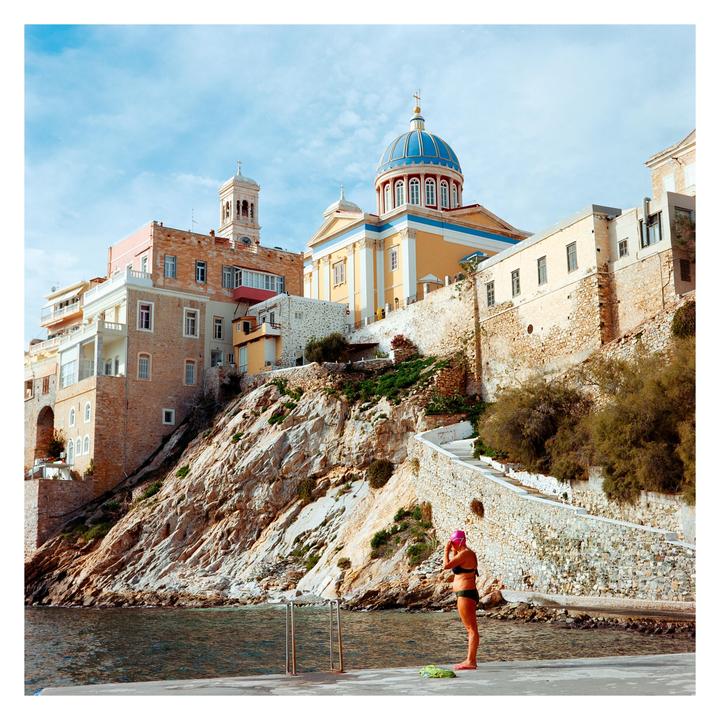 A woman in a swimsuit and swim cap stands on a concrete pier next to a body of water, with buildings and a large church on a rocky hillside in the background.