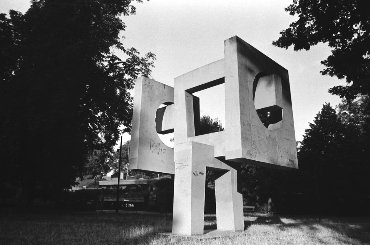 A large concrete sculpture with geometric cutouts stands in a grassy area surrounded by trees.