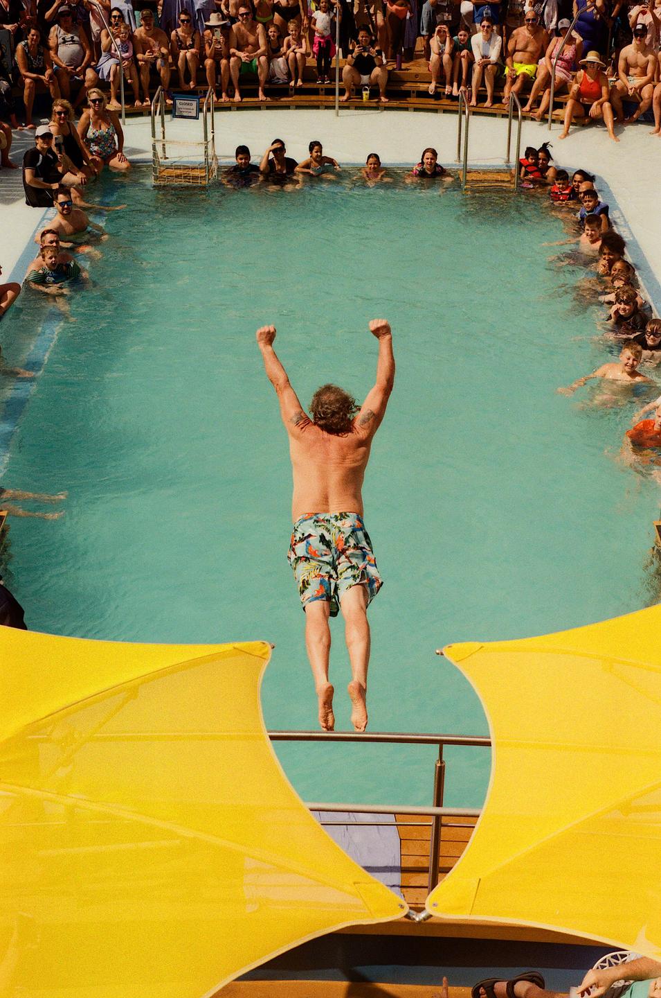 A man jumps into a swimming pool while a crowd watches from the edge and from a deck above.