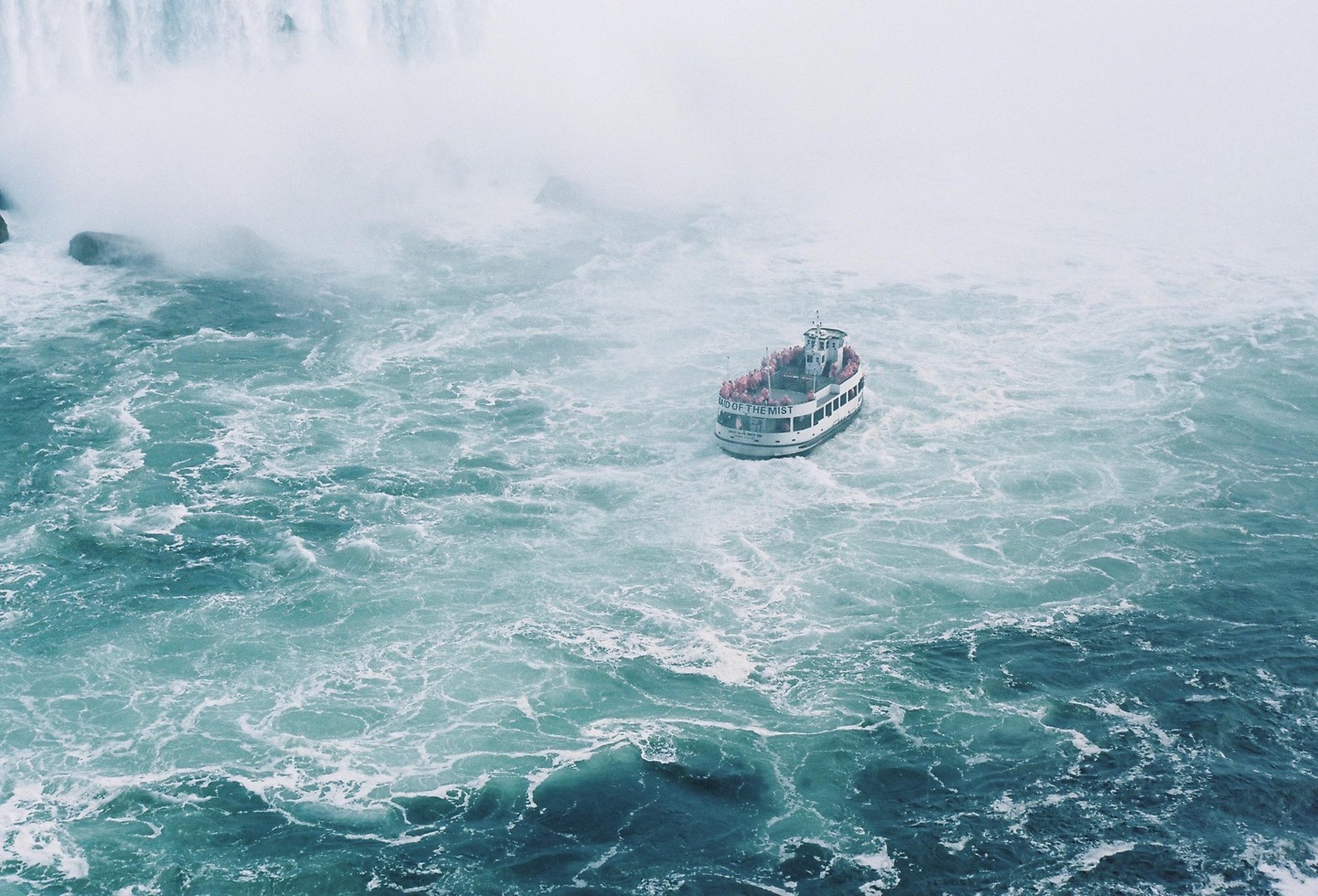 A boat full of people in red ponchos navigates the turbulent water at the base of a waterfall.
