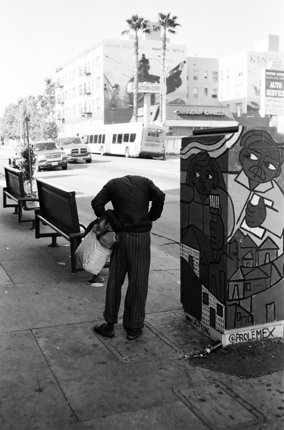 A person with no head stands on a sidewalk next to a painted utility box and two empty benches.