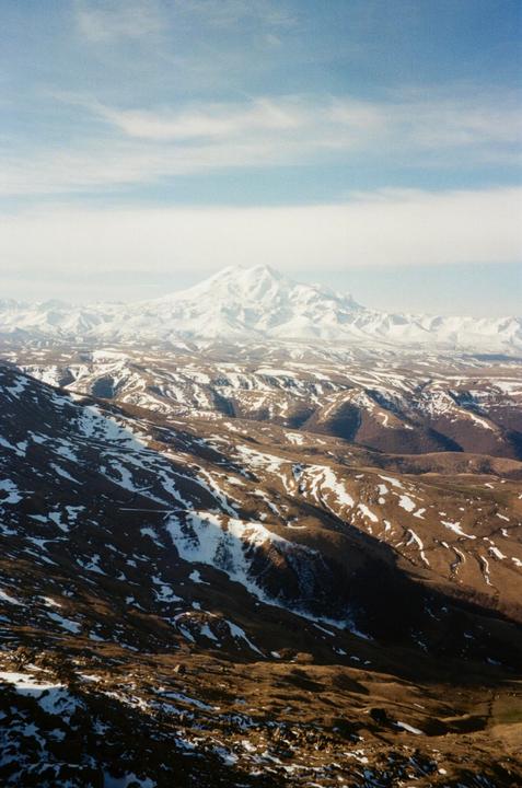 A vast mountain landscape shows snow-capped peaks under a partly cloudy sky.