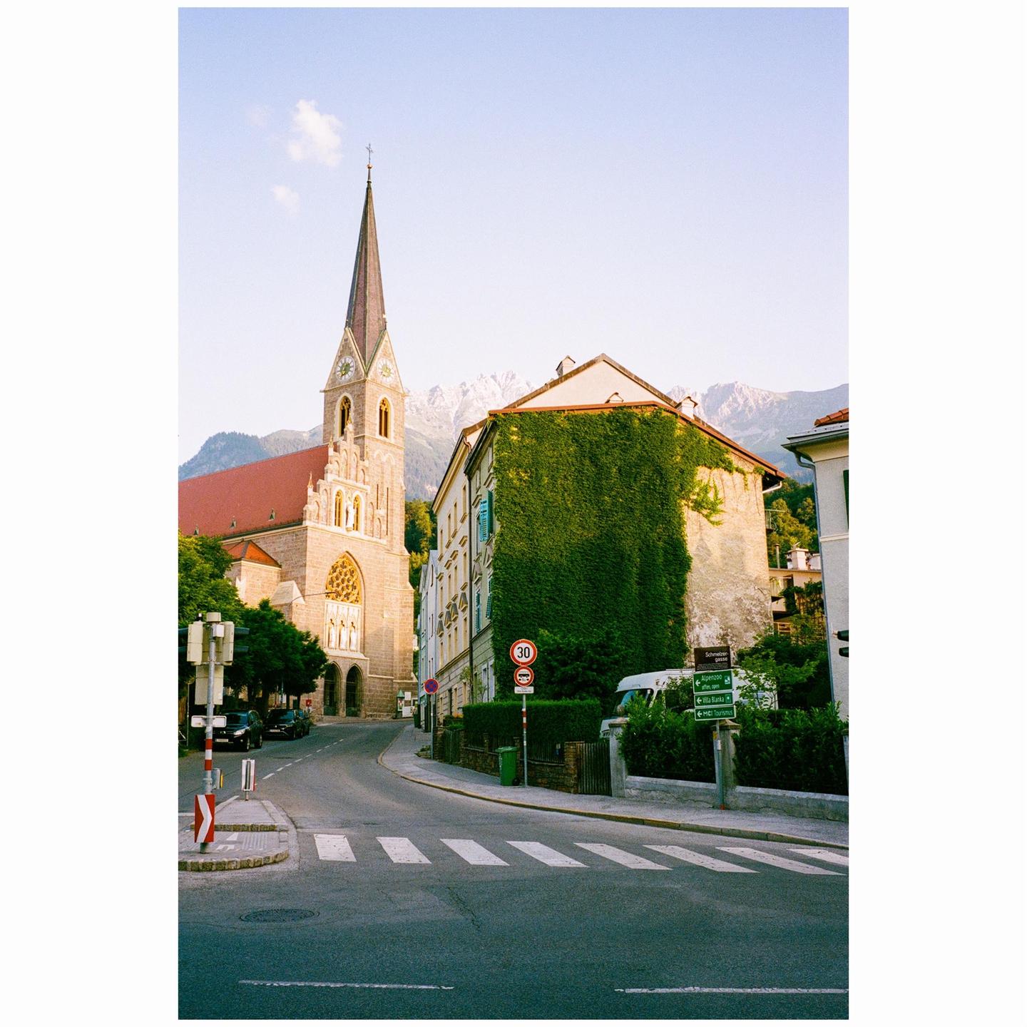 A street curves past a church with a tall spire and a building covered in ivy, with mountains in the background.