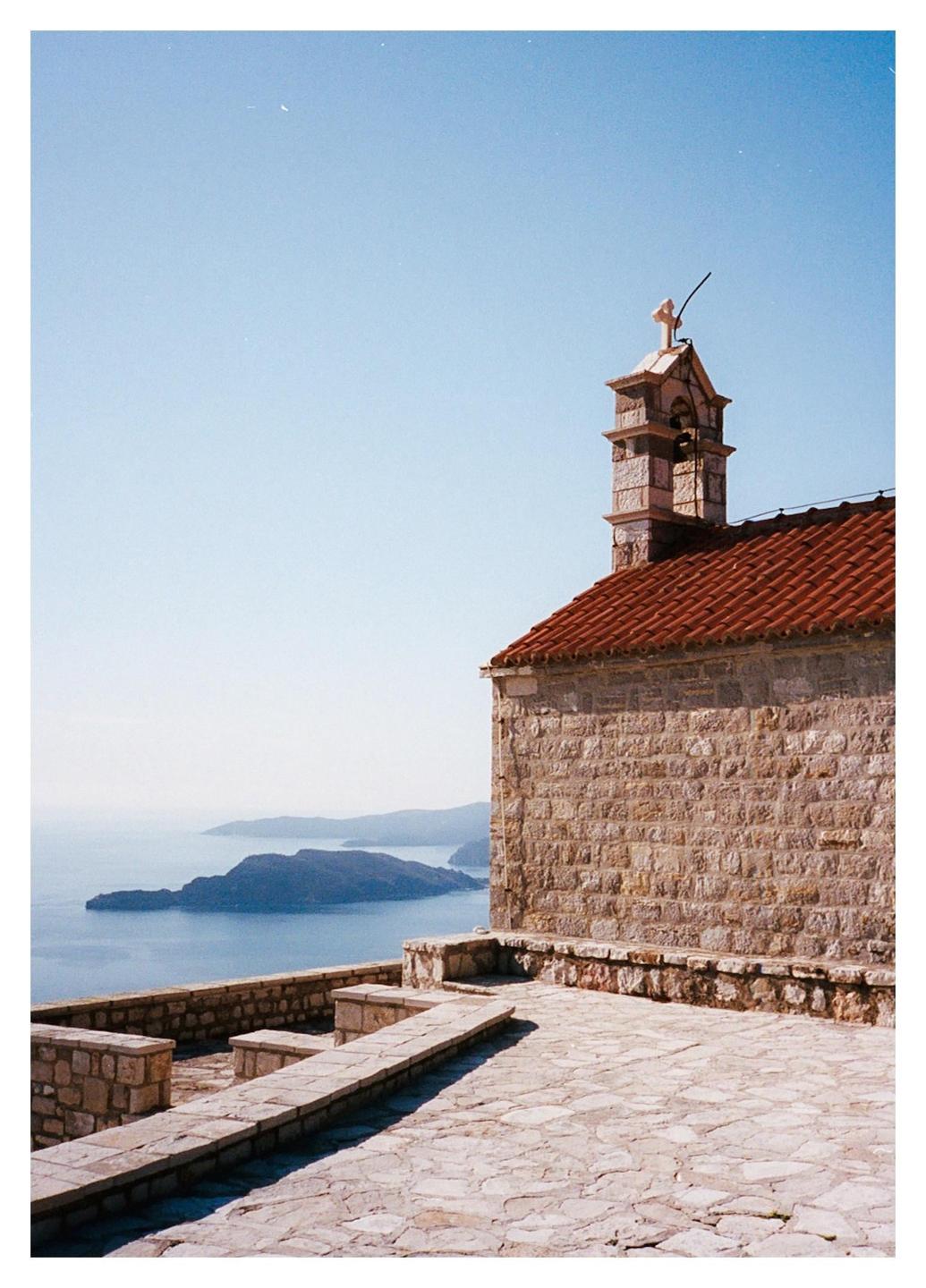 A stone building with a red tile roof and bell tower overlooks a body of water with islands in the distance.