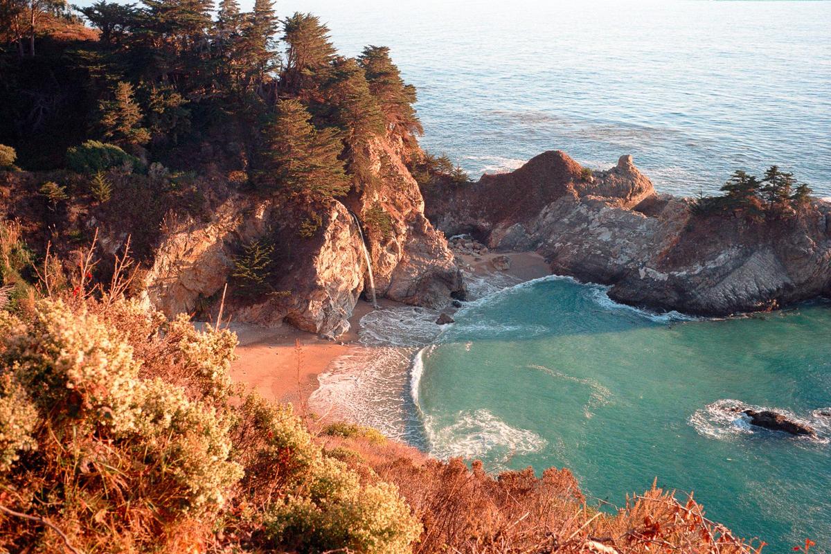 A waterfall cascades onto a sandy beach next to a turquoise ocean, surrounded by cliffs and pine trees.