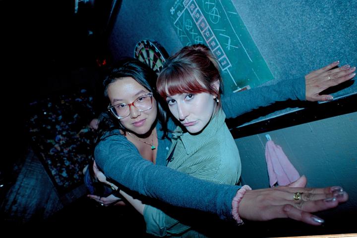 Two women are embracing each other in a dimly lit room with a dartboard and a scoreboard on the wall.
