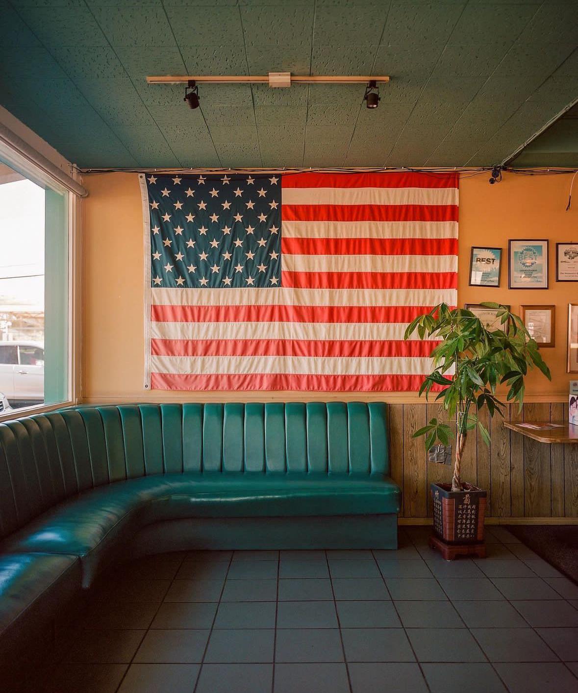 An American flag hangs above a green booth in a diner with a potted plant on the right.