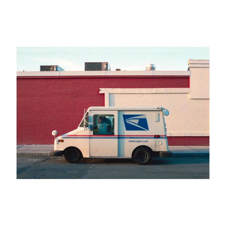 A white USPS mail truck is parked on a street next to a building with a red brick wall.