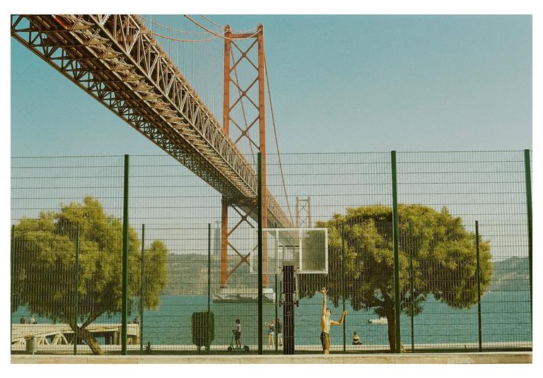 A boy plays basketball on a court next to a body of water, with a large bridge overhead.