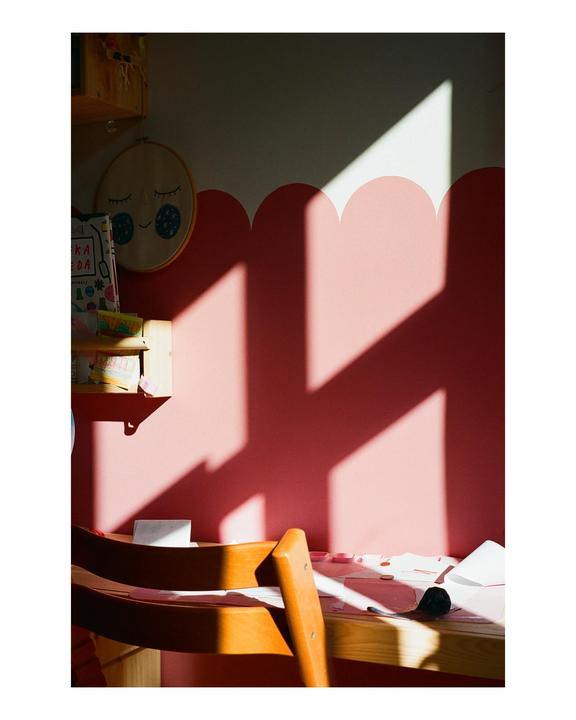 Sunlight casts geometric shadows across a pink and white wall above a wooden desk and chair.