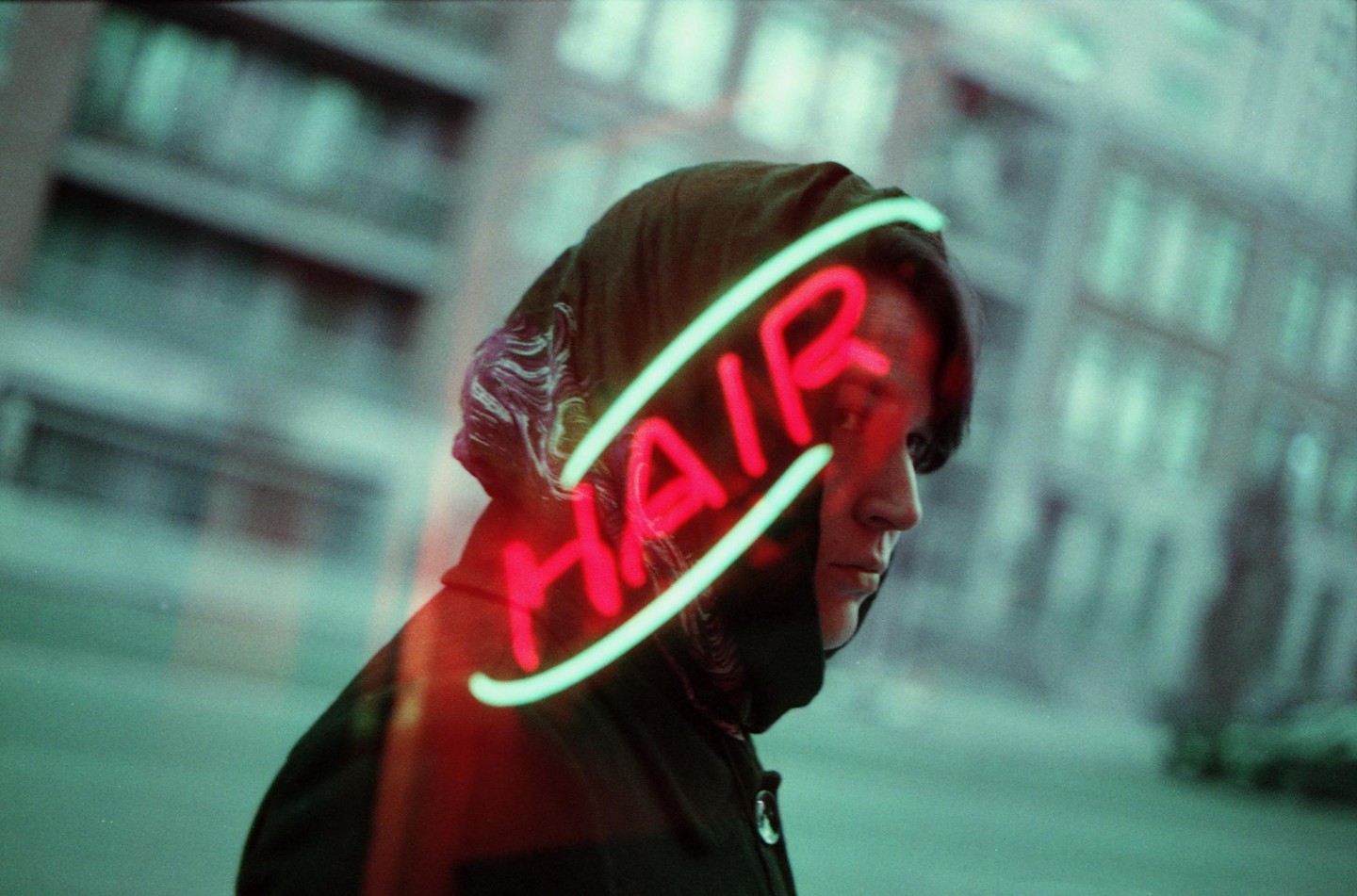 A person with a dark hood looks to the right, with a neon sign that says "HAIR" reflected across their face.