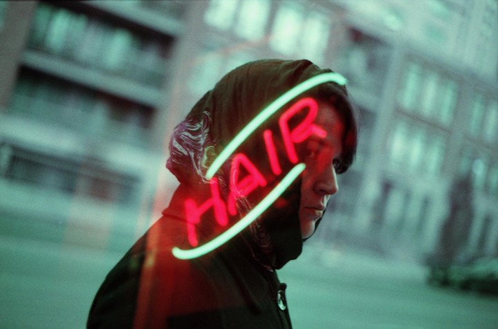 A person with a dark hood looks to the right, with a neon sign that says "HAIR" reflected across their face.