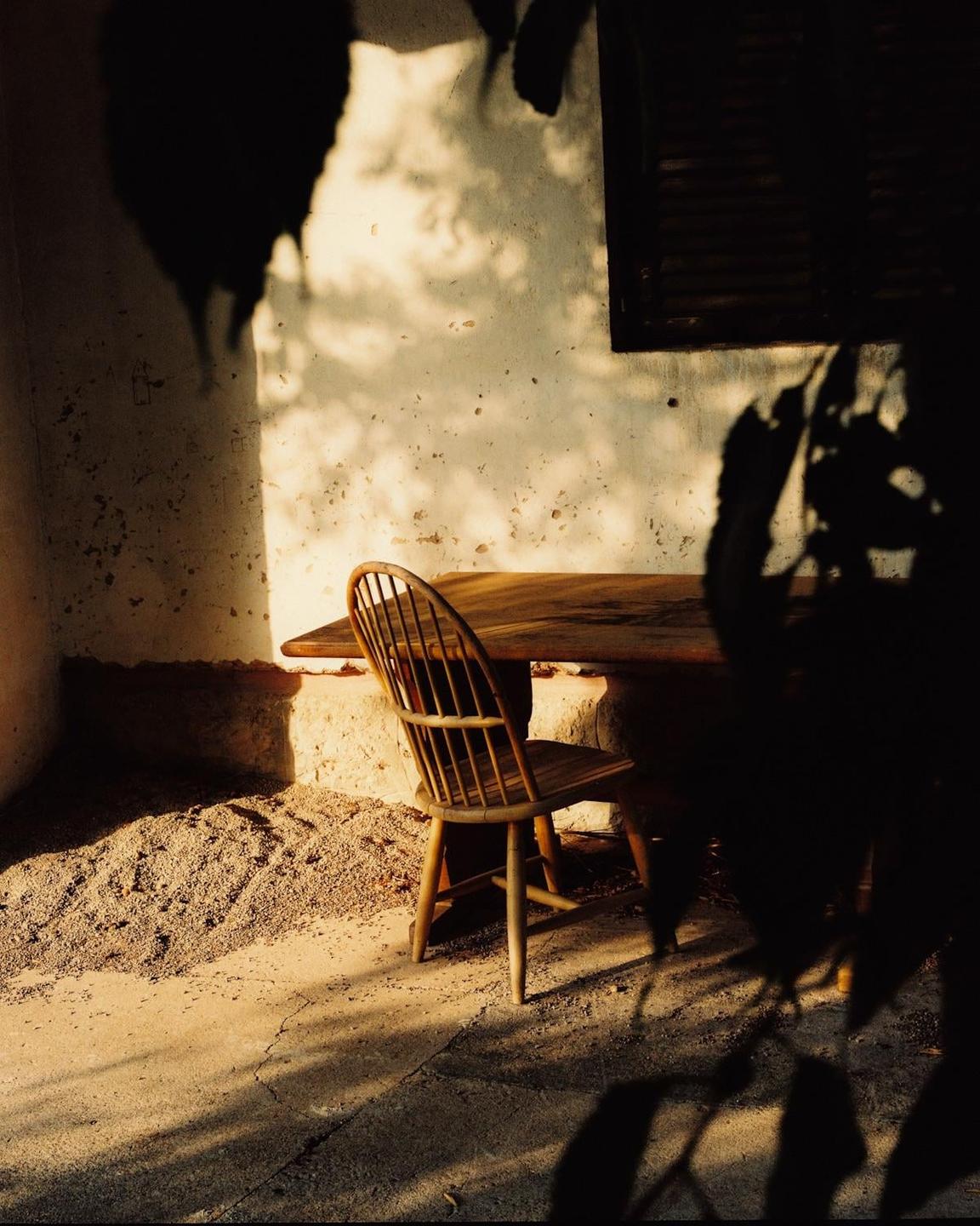 A wooden chair and table are illuminated by dappled sunlight on a textured wall and ground.
