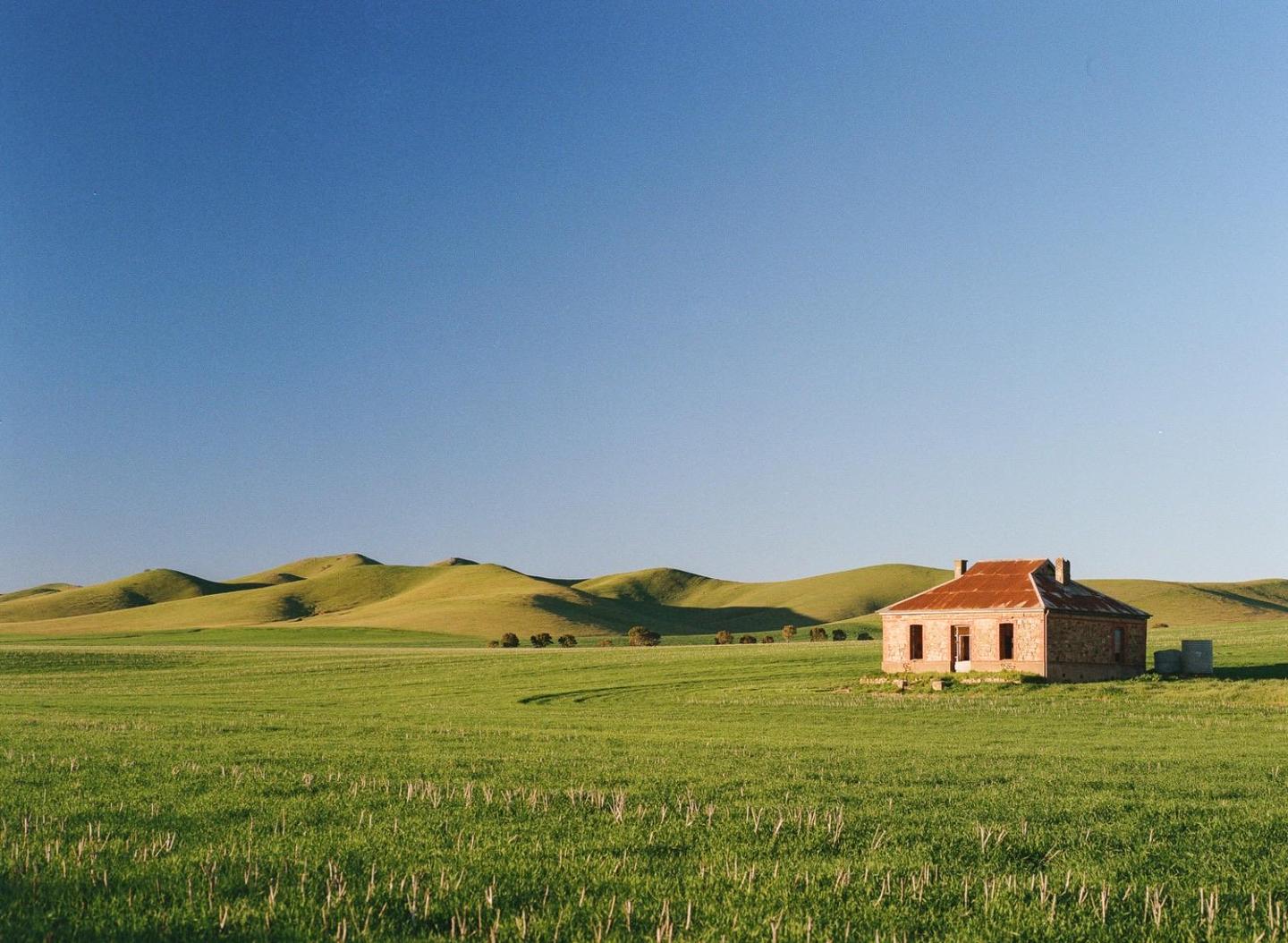An abandoned stone house with a rusty roof stands in a green field under a clear blue sky, with rolling hills in the background.