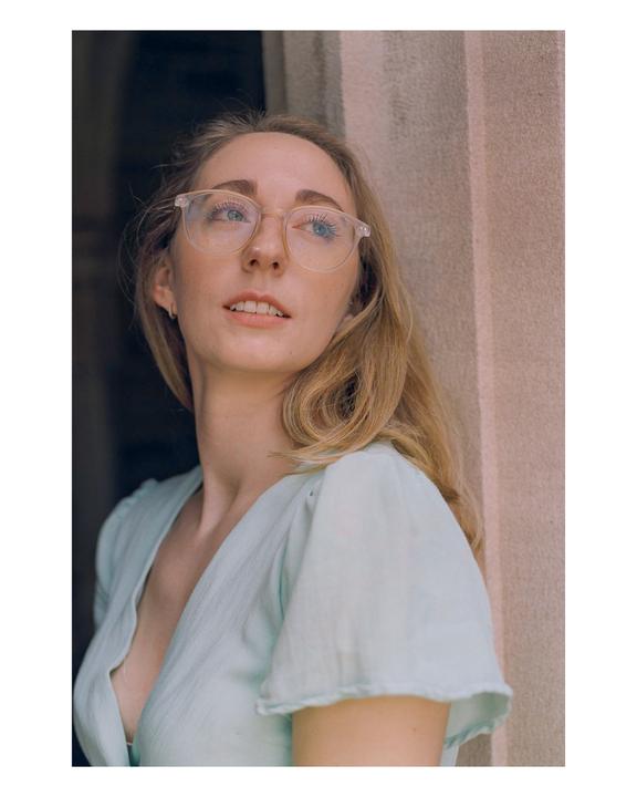 A woman with long blonde hair and glasses looks up and to the right, wearing a light blue top.
