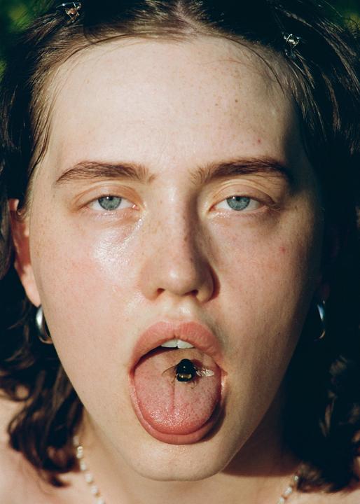 A woman with a bumblebee on her tongue stares directly at the viewer.