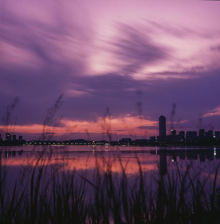 A long exposure captures the movement of clouds over a city skyline reflected in a lake at dusk, with tall grass in the foreground.