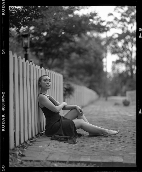 A woman with a shaved head sits on a brick path next to a white picket fence.