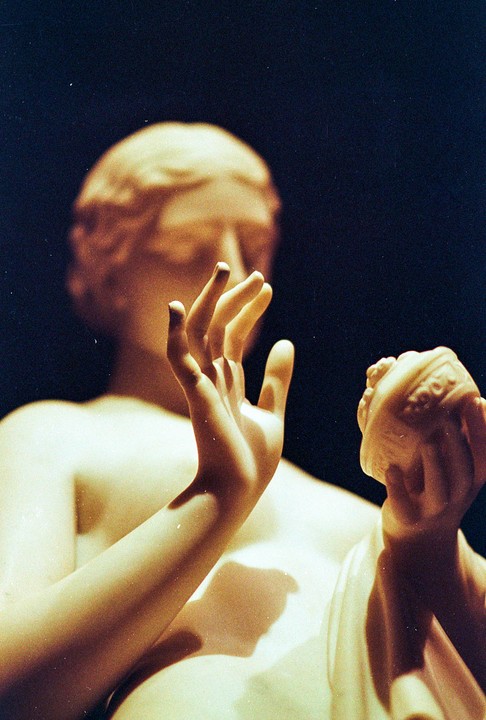 A close-up of a classical sculpture shows a hand reaching out and another hand holding a shell.