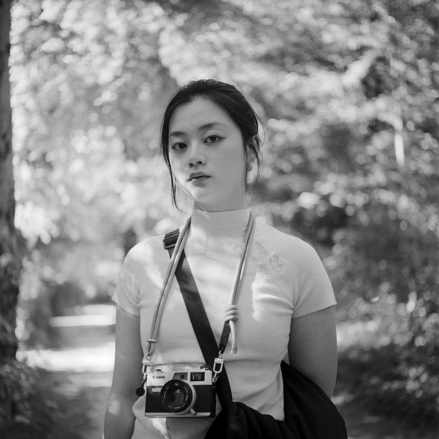A woman wearing a turtleneck and carrying a camera stands in a forest.