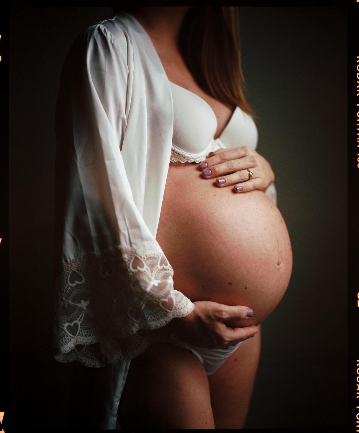 A pregnant woman in a white robe and bra holds her belly.