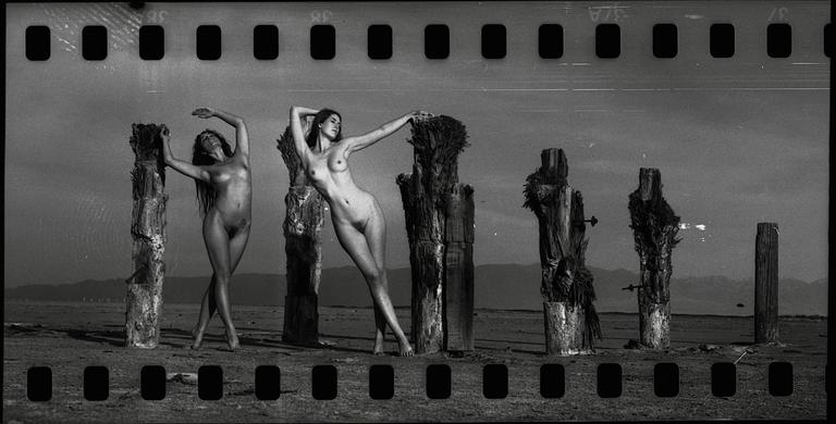 Two women pose nude next to weathered wooden posts in a barren landscape with mountains in the distance.