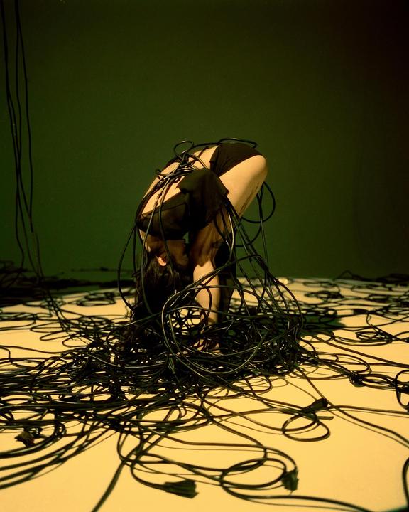 A woman is curled up on the floor, entangled in a mess of black cables.