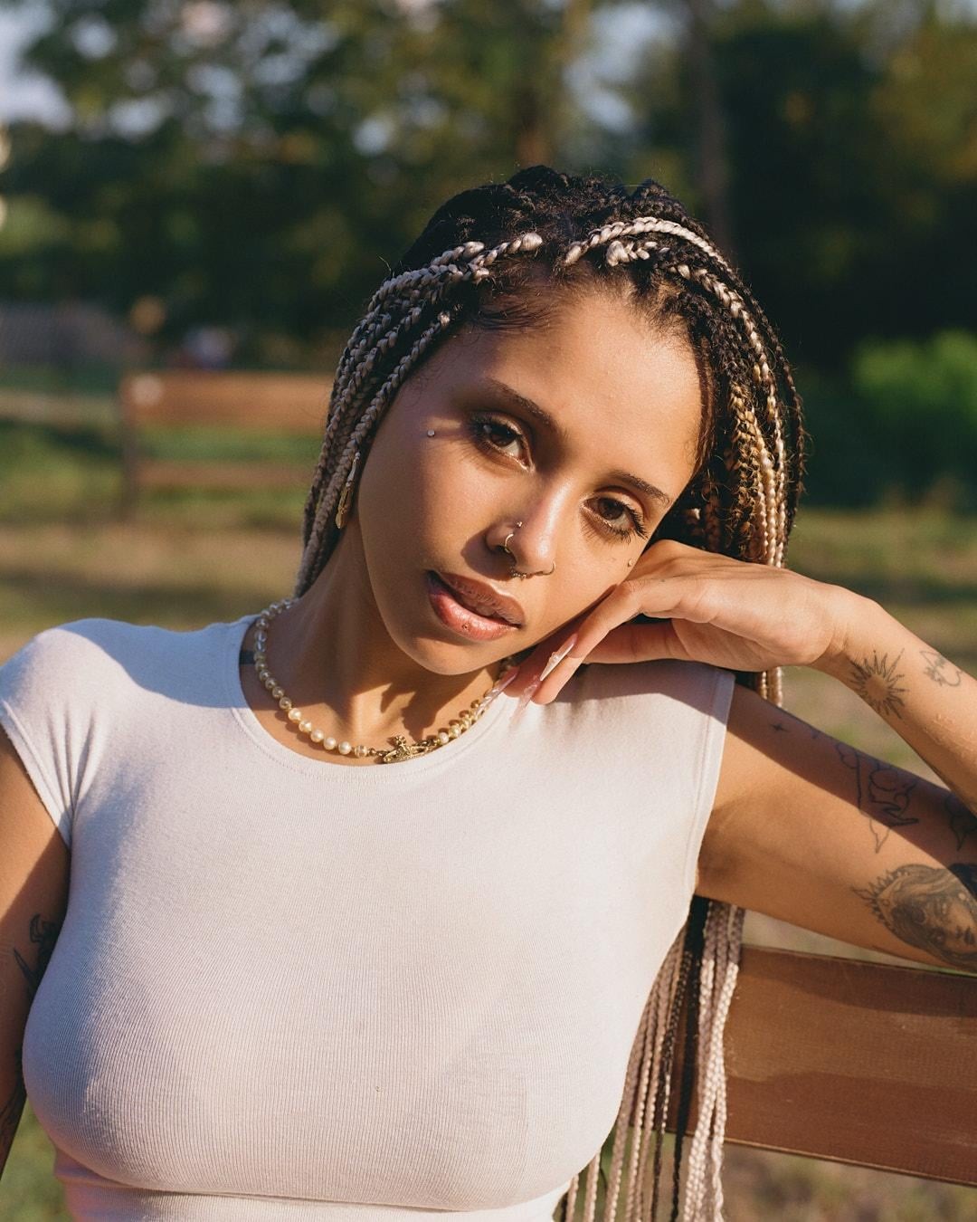 A woman with braided hair and tattoos on her arm leans on a wooden bench in a park.