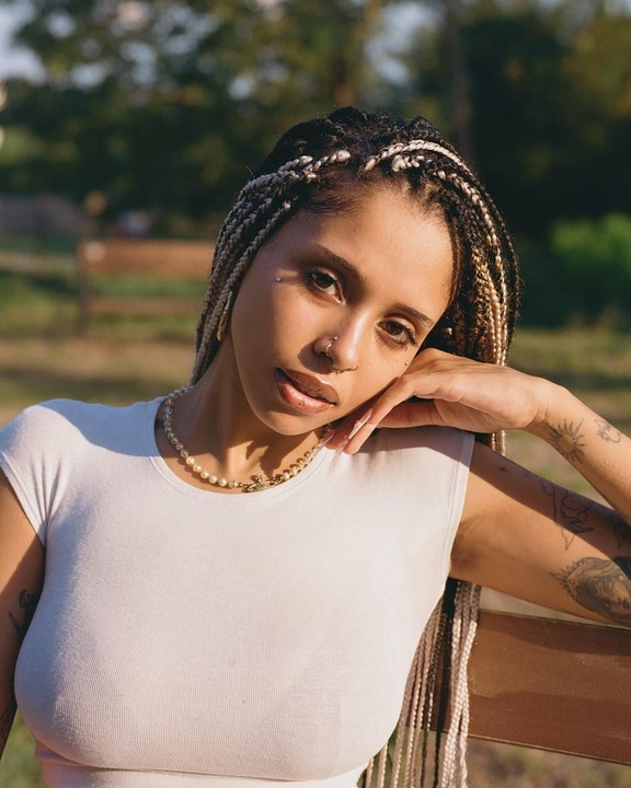 A woman with braided hair and tattoos on her arm leans on a wooden bench in a park.