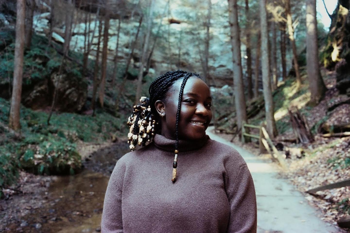 A woman with braided hair and beads smiles while standing in a forest with a path and stream.