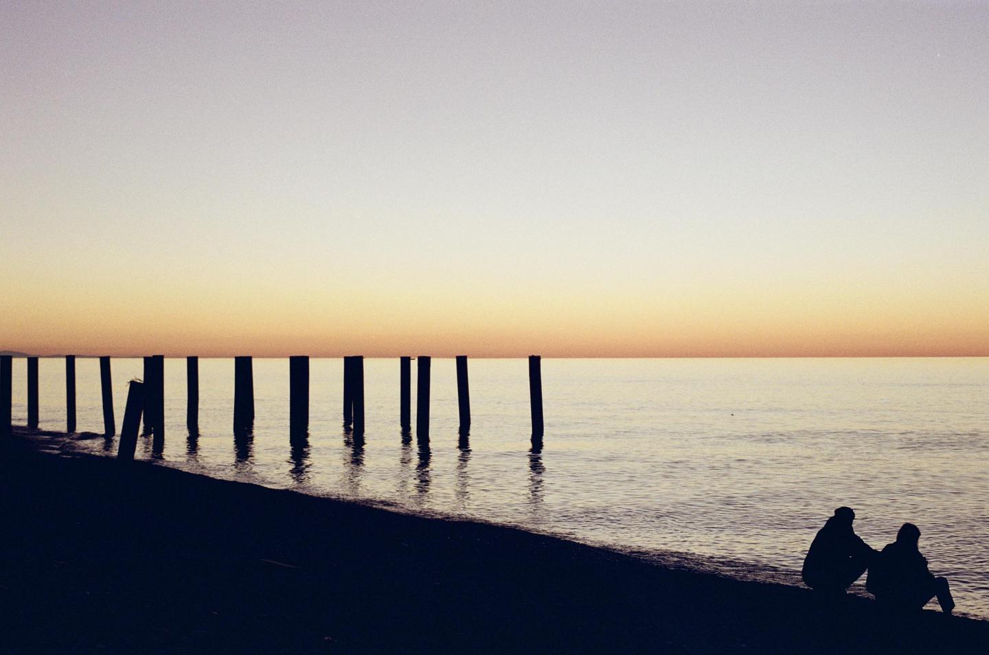 Two people sit on a beach at dusk with a row of pier pilings extending into the water.