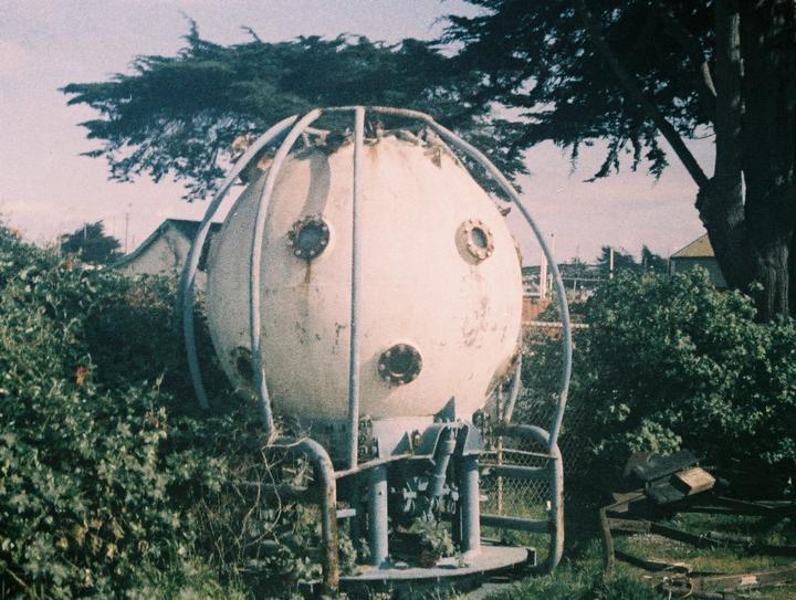 A large, spherical, white diving bell with portholes is surrounded by green bushes and trees.