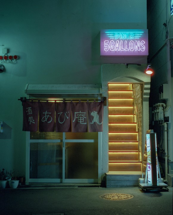 A neon sign for "5 GALLONS DINER" hangs above an illuminated staircase next to a shop with a red noren.