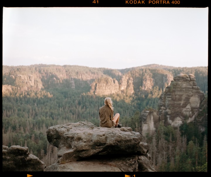 A woman sits on a large rock formation, looking out at a valley filled with trees and other rock formations.