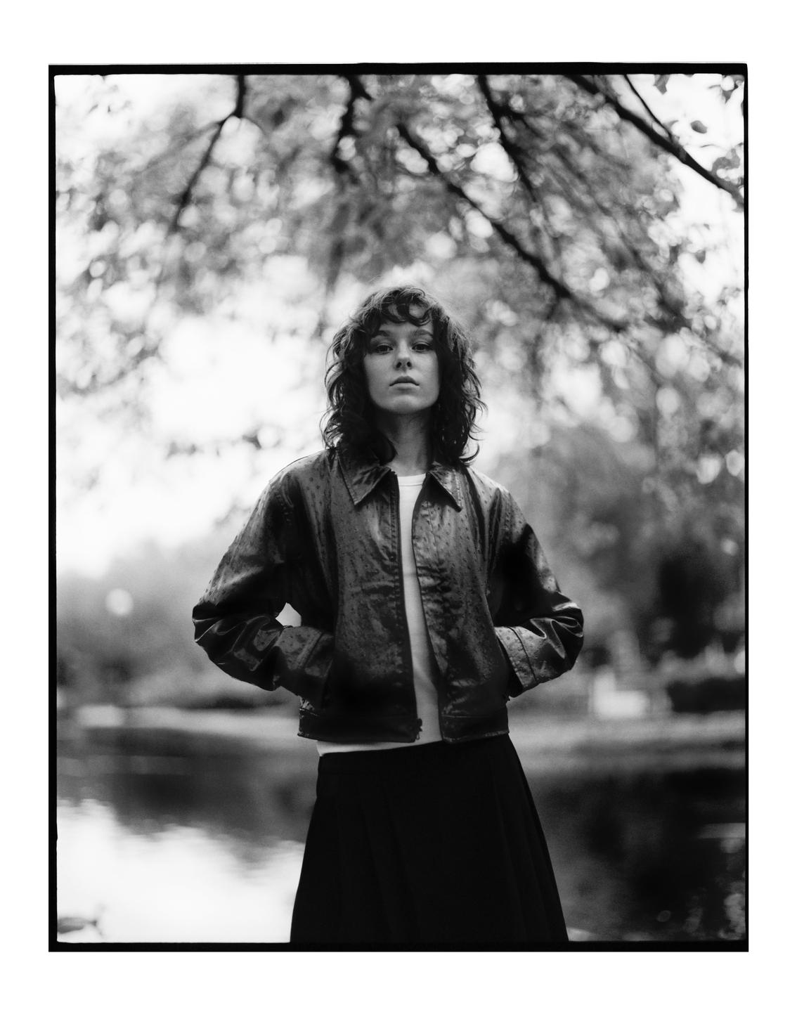 A woman with curly hair stands with her hands in her pockets, with a body of water and trees in the background.