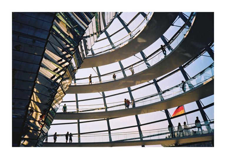 People walk along a spiral ramp inside a glass dome with a German flag visible in the background.