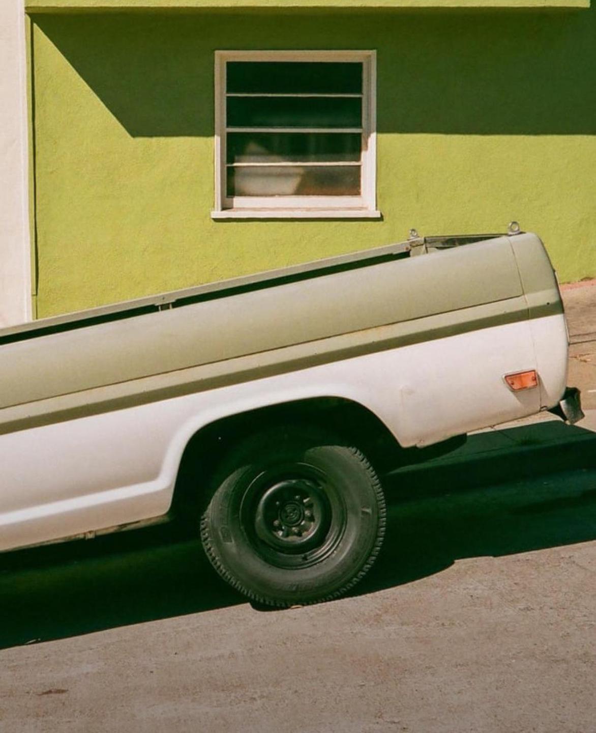A white and green pickup truck is parked on a street next to a lime green building with a window.