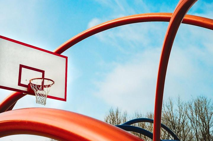 A basketball hoop and backboard are visible against a blue sky with abstract orange and blue structures in the foreground.