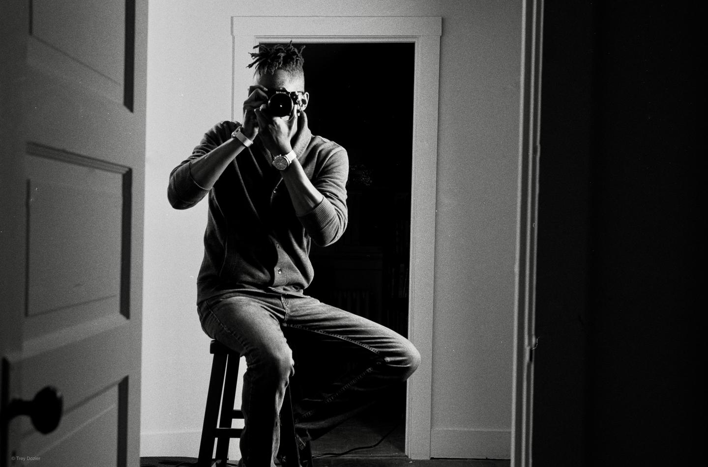 A man with dreadlocks sits on a stool and photographs his reflection in a mirror.