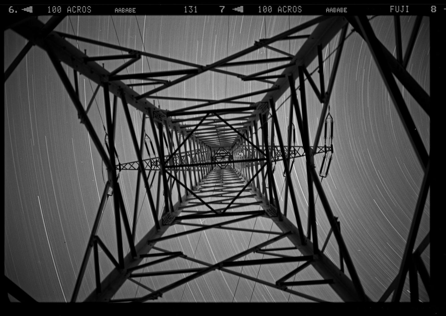 A low-angle view of a metal electricity pylon shows star trails in the night sky.