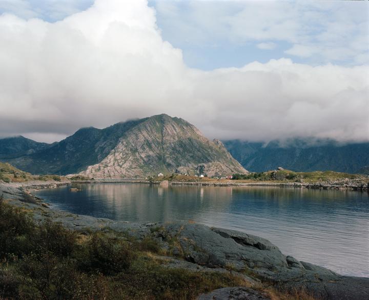 A body of water reflects a large mountain under a cloudy sky.