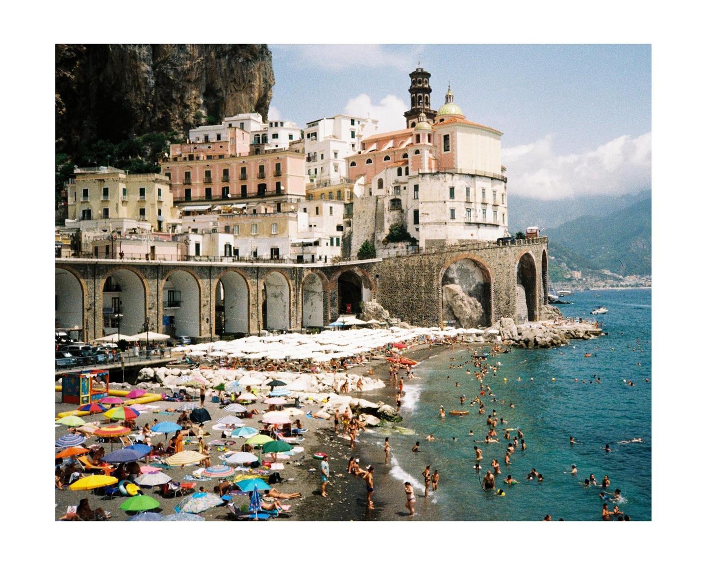 A crowded beach with colorful umbrellas is visible next to a town built into a cliffside.