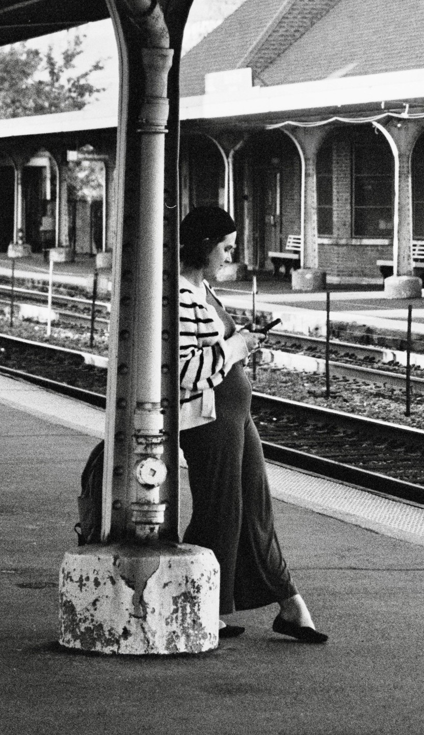 A pregnant woman stands on a train platform, leaning against a pillar and looking at her phone.
