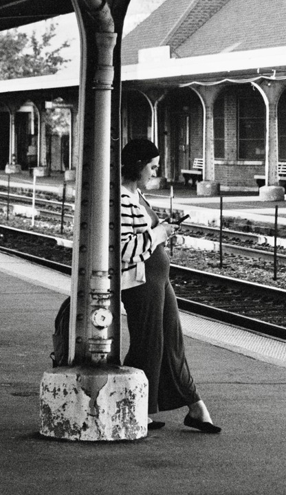 A pregnant woman stands on a train platform, leaning against a pillar and looking at her phone.