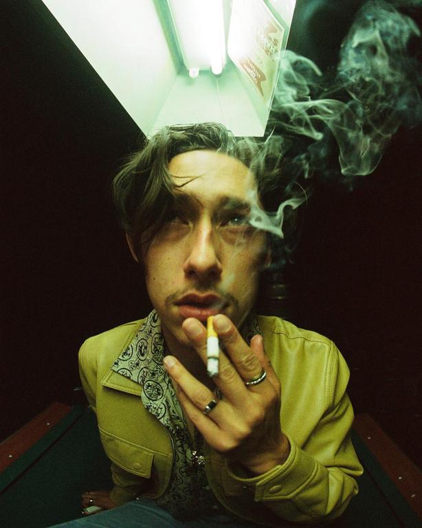 A man with a mustache and rings on his fingers smokes a cigarette, with smoke swirling around his head, under a fluorescent light.