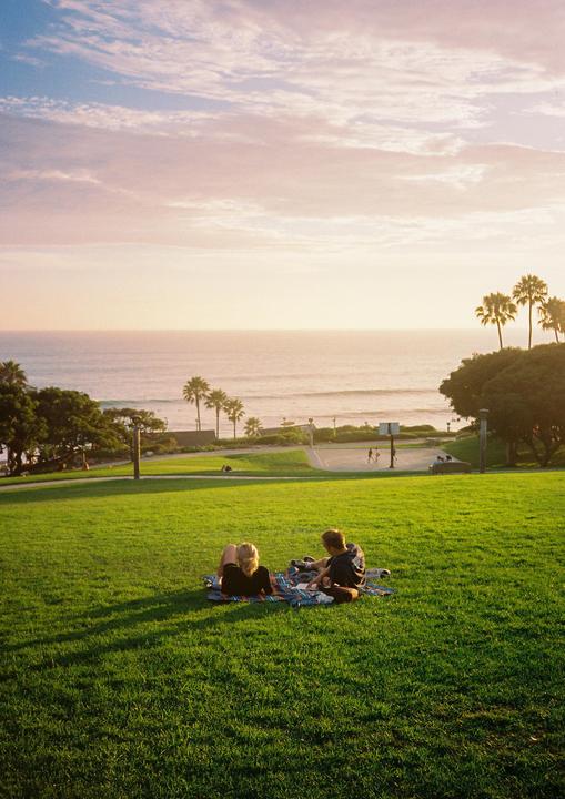 A couple sits on a blanket in a grassy park overlooking the ocean at sunset.